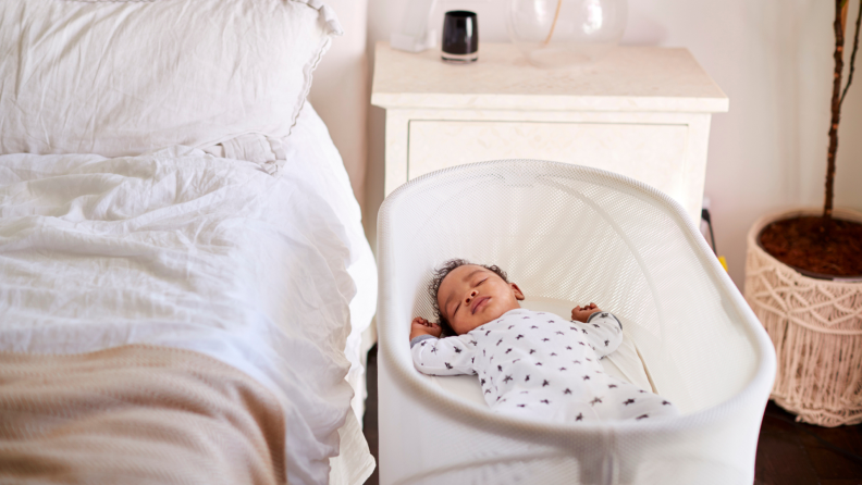 Small infant sleeping inside of bedside bassinet next to bed in bedroom.