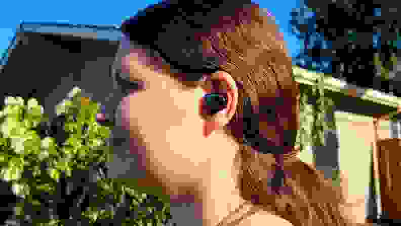 The mirrored black Master & Dynamic MW08 earbud sits in the ear of a pretty young brunette woman with the backdrop of lush greenery and a tan house.