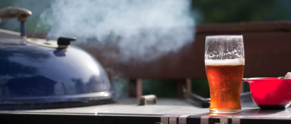 A pint glass of beer sits next to a smokey grill