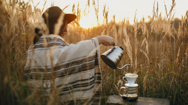 A person with a hat and blanket is pouring water into a pour-over dripper to make coffee in the fields.
