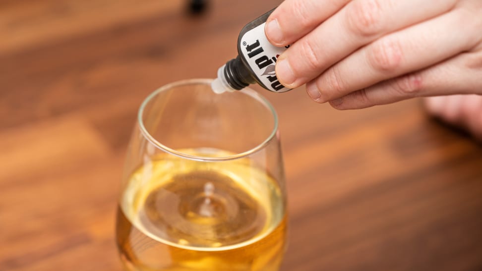 A hand holds a bottle of Drop It wine drops poised above a glass of white wine