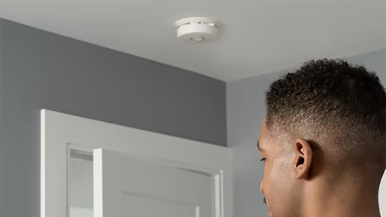 Person standing in room with circular smoke detector mounted on ceiling.