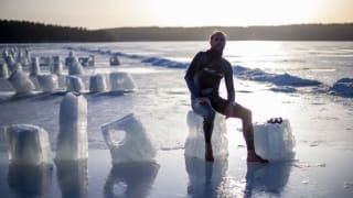 A person in a wetsuit sits atop a block of ice on the beach.