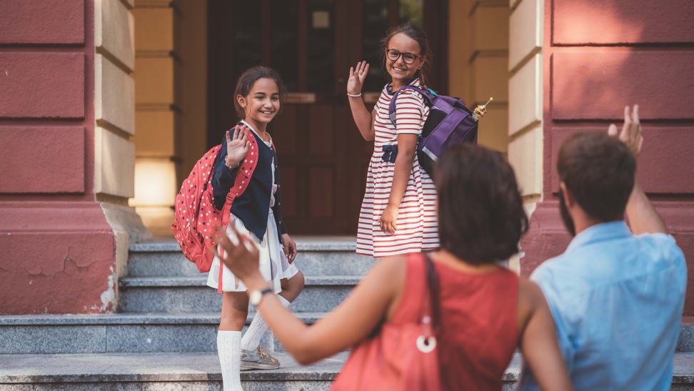 Two young girls on the steps of a school waving to their parents in the foreground