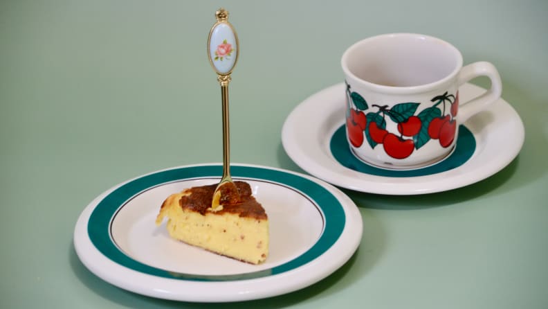 A slice of Basque cheesecake is served nearly on a white-and-green plate beside a mug with cherries painted on it.