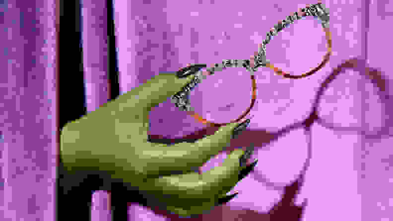Witches hand holding Halloween-themed eyewear on purple background.