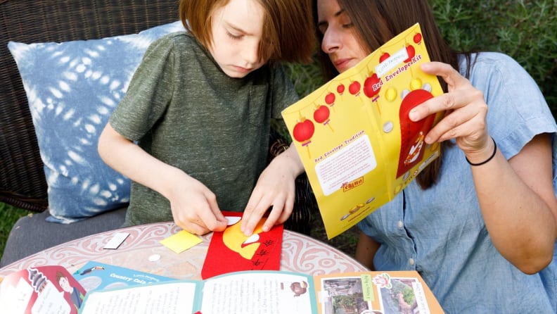 A child works on a craft making a Chinese money envelope, while his mother looks on