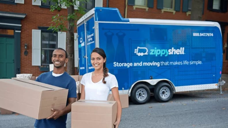 A couple smiling for the camera before continuing to place boxes on the ground behind a portable storage box in front of an apartment