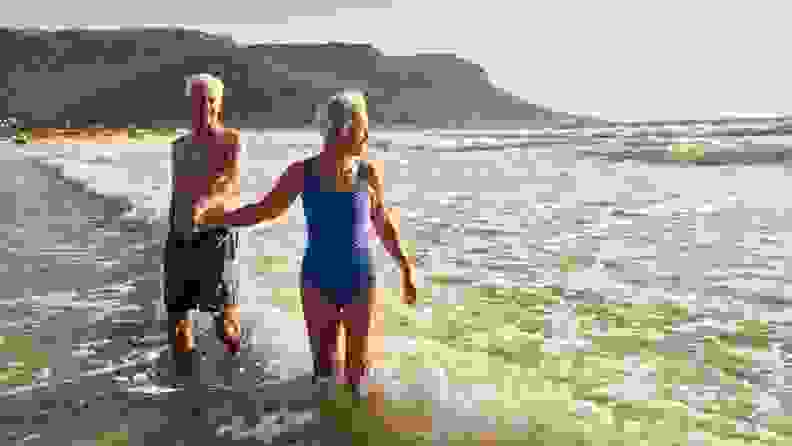 An elderly couple wearing bathing suits walks out of the ocean after a swim