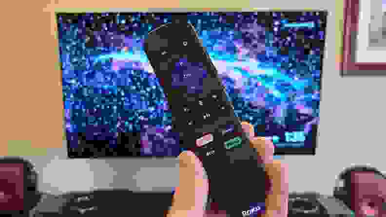 The Roku Streaming Stick 4K's black remote is held before an OLED display with stars strewn across the background, bearing multiple streaming and control keys.