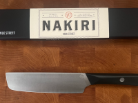 A Milk Street Nakiri Knife underneath the box packaging on top of a wooden cutting board.