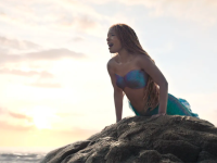 An image of Halle Bailey as Ariel the mermaid in "The Little Mermaid;" in the image, she is perched on a rock against a sunlit ocean sky.