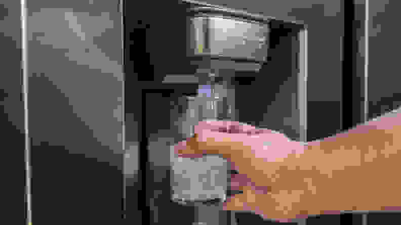 A hand reaches into frame holding a glass up to the Samsung RS27T5200SR's through-the-door dispenser.