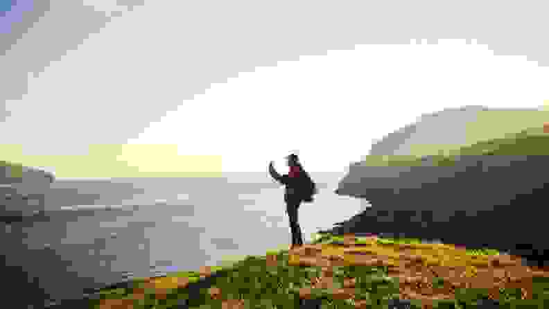 A hiker with a backpack stands on a cliffside to snap a photo of the sea