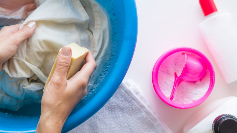 A person's hands scrubbing fabric in a bucket filled with sudsy water.