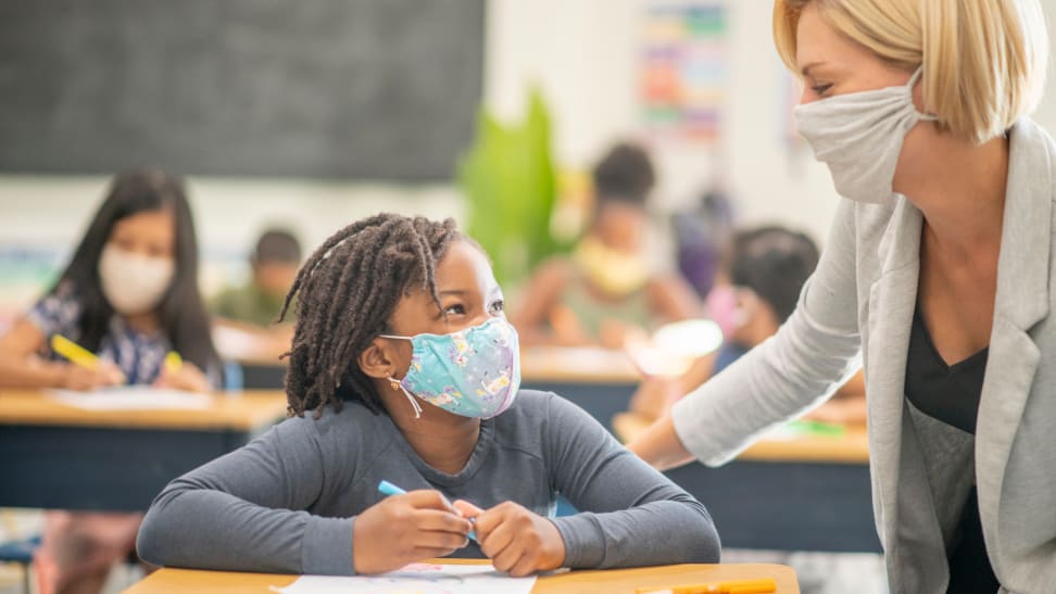Woman wearing face mask standing over young girl who is also wearing a face mask, while holding pencil and sitting at a desk in a classroom with other students