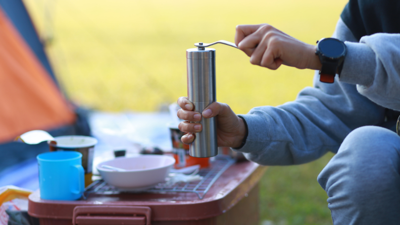 A person is using a hand-crank grinder at what appears to be a camping site.