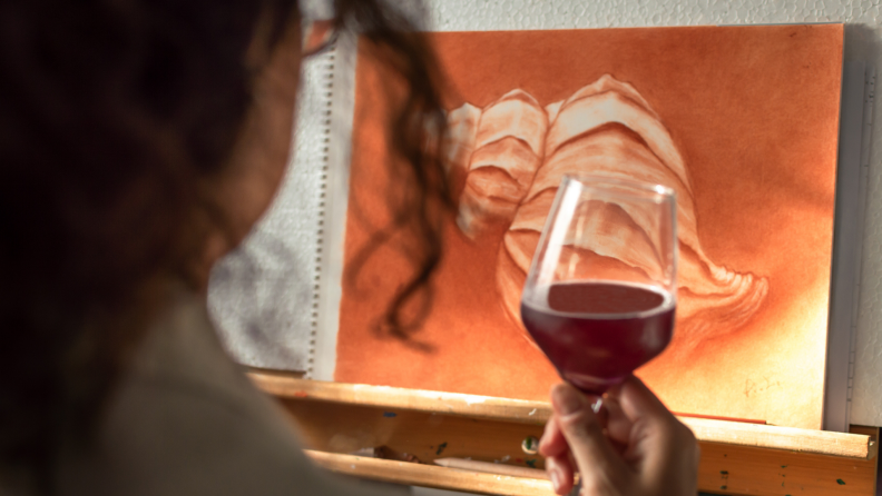 Person painting a picture while drinking a red glass of wine