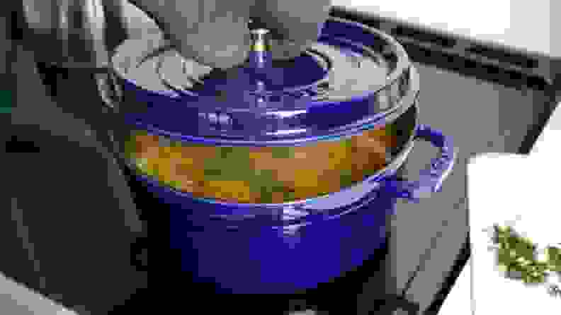 A chef lifting the lid on a Staub Dutch oven, revealing soup inside.