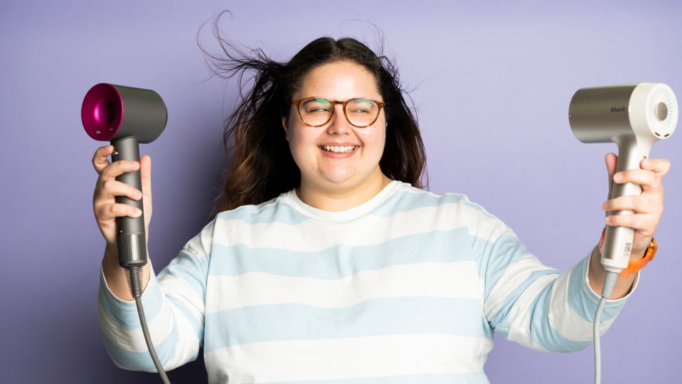 A person with long hair wearing a blue and white striped shirt stands against a purple background and points two hair dryers at their hair.