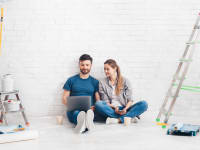 Two people sitting against a white wall surrounded by home improvements supplies.