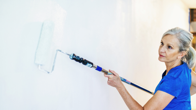 Senior woman wears her hair back in a ponytail as she uses a extended roller brush to paint wall.