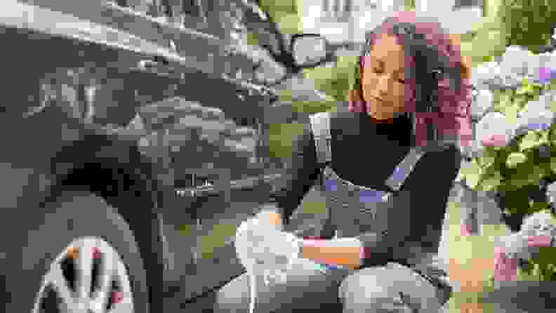 A young woman kneels by the rear wheel of her car to clean it with a sponge