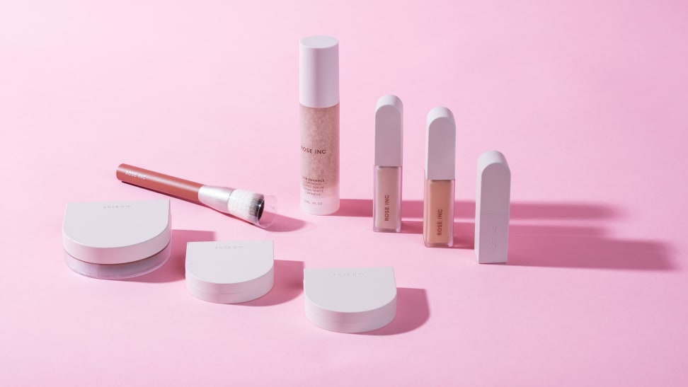 Rose Inc. beauty lip and cheek blushes, lipstick and concealer tubes and makeup brush against pink background.