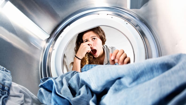 A shot from the back of a front-loader's drum shows a woman peering in at her laundry, pinching her nose in exaggerated disgust.