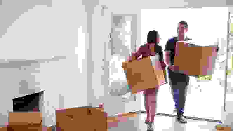 A man and woman walk into their new home holding cardboard moving boxes