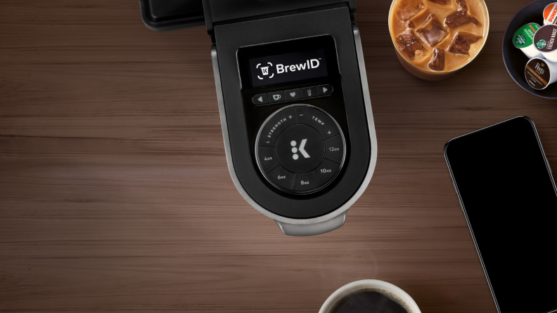 On a brown wooden table, there's a Keurig K-Supreme Plus Smart  pod coffee maker with BrewID technology and a glass of iced coffee next to it.