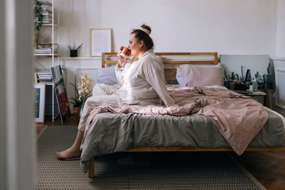 Plus-size woman sitting on edge of bed drinking coffee wearing PJs