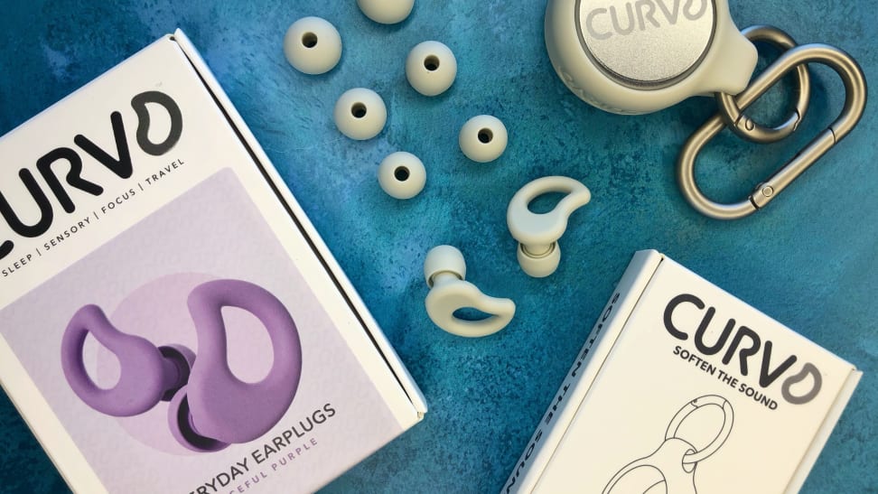 A box of purple CURVD ear plugs, as well as white CURVD earplugs laid out on a blue fabric.