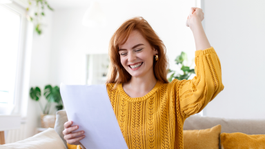 A woman in a yellow sweater smiles and raises her fist while reading a document, celebrating good news.