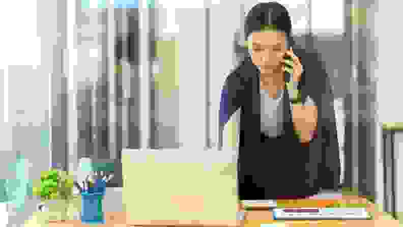 Woman dressed in professional attire in front of laptop, using phone and working.