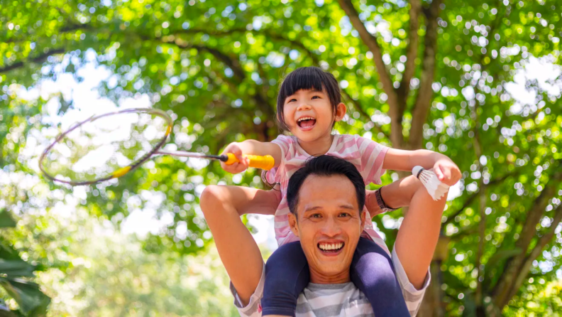 A parent carries their child over their shoulders outside.