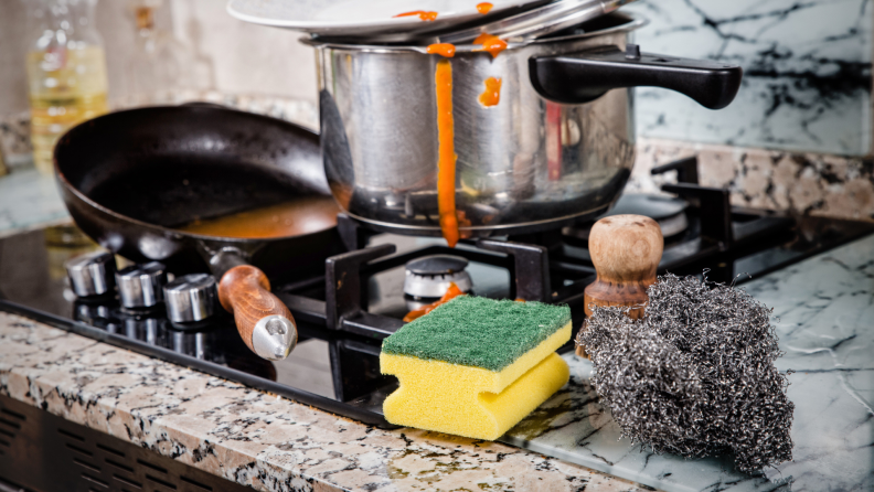 Dirty dishes stacked up on a stovetop with a sponge and steel wool ball on the counter