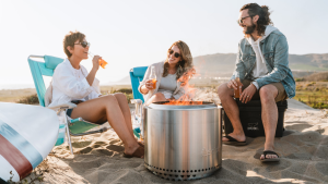 A group of people on a beach sitting on folding chairs surrounding a Solo Stove.