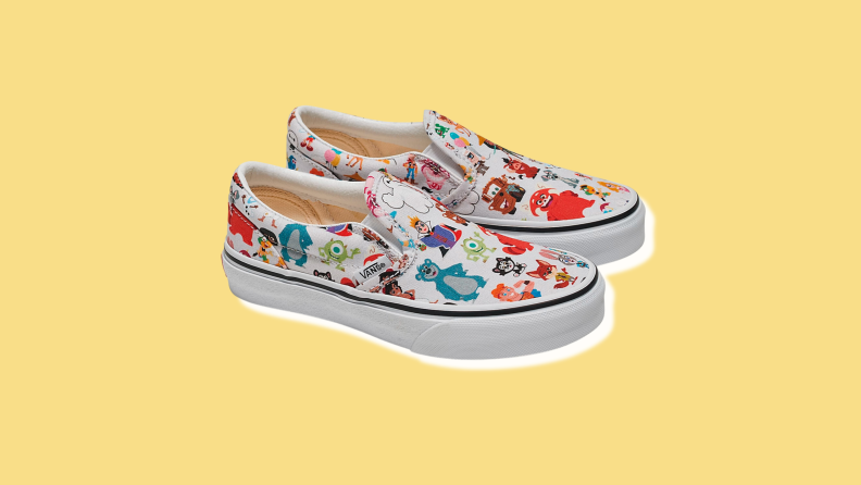 A pair of white slip-on Vans sneakers with an all-over pattern of Disney cartoon characters.