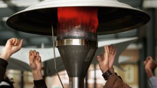 A small gathering of folks warm their hands around a glowing patio heater.
