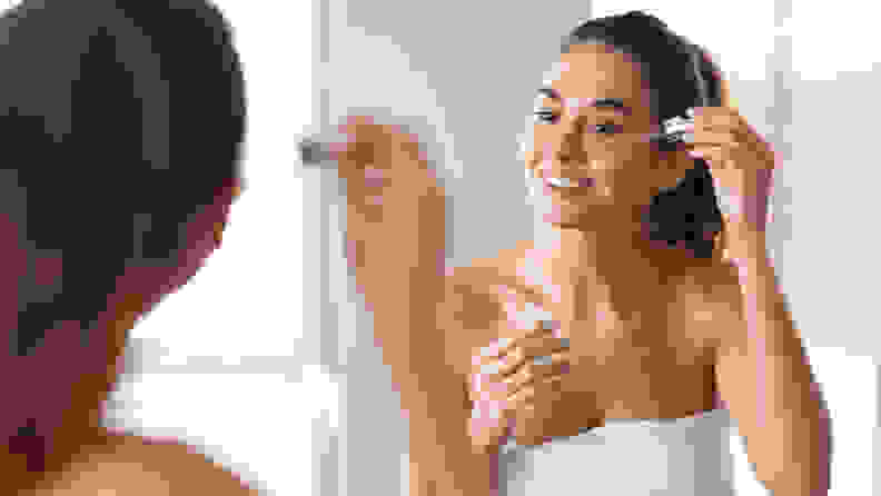 A person looks into a mirror and applies a dropper of serum onto their skin while wearing a bath towel.