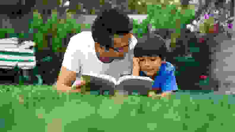 A dad looks at a book with his son as they lay on the grass.