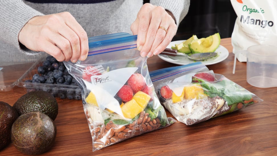 Person closing a freezer bag of smoothie ingredients on a wood counter with other bag and ingredients around it.