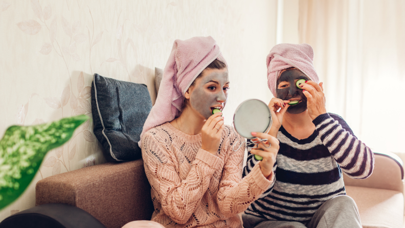Adult daughter and mom with face masks and towels on their hair putting cucumbers on their eyes