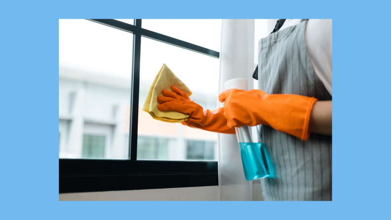Person with rubber gloves and apron using spray bottle and microfiber towel to clean windows.