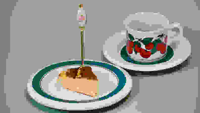 A slice of Basque cheesecake is served nearly on a white-and-green plate beside a mug with cherries painted on it.