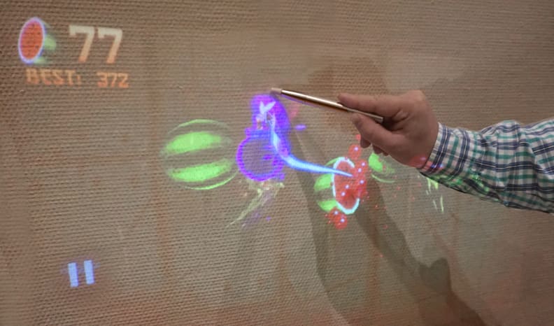 A demonstrator plays Fruit Ninja on a wall in the exhibit hall using the TouchPico.