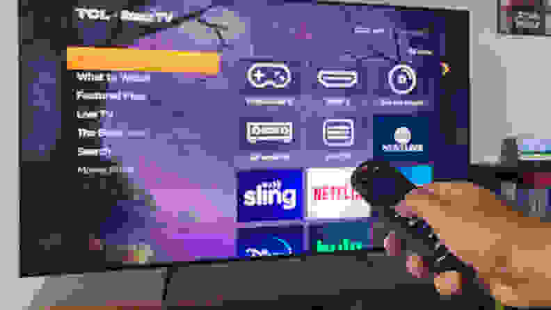 A hand holding a Roku remote pointing at a TCL 5-Series TV displaying the Roku OS home screen.