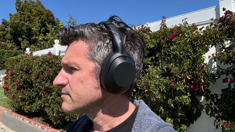 A man wearing the Sony Ult Wear headphones outside on a sidewalk with bushes behind him.