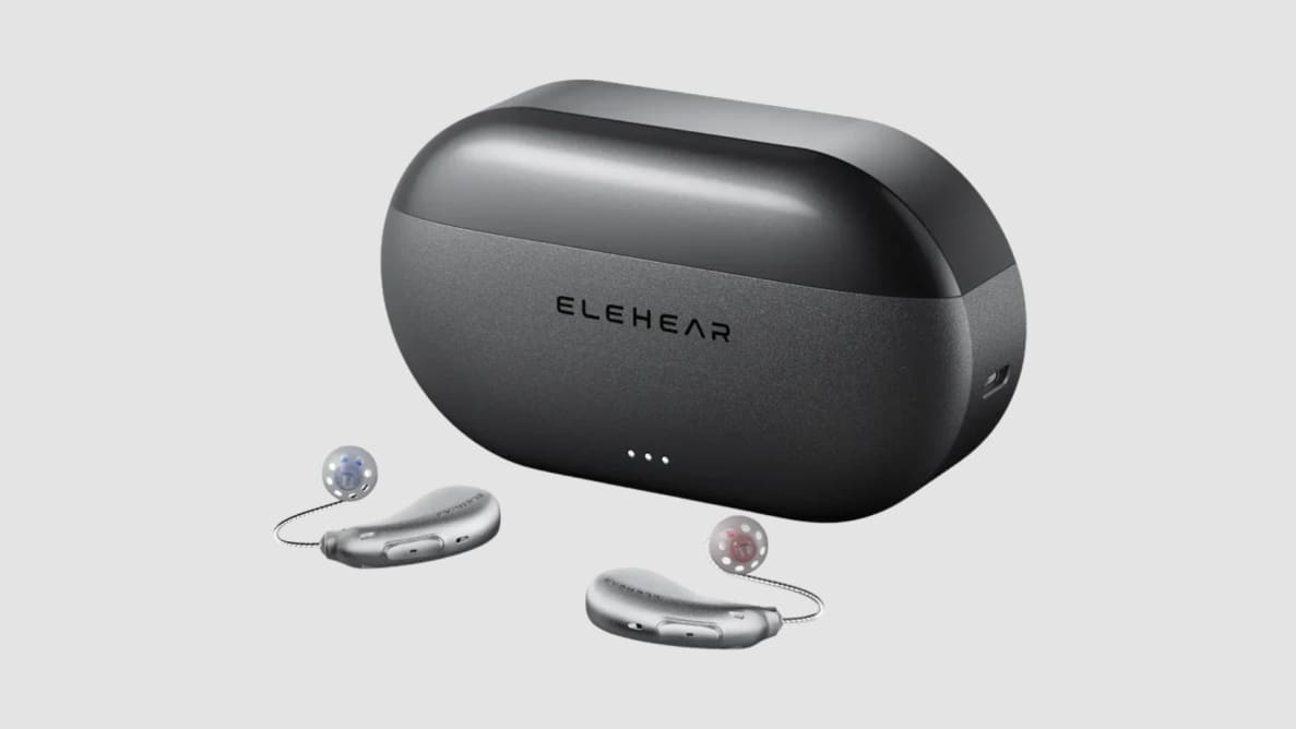 A black oval charging case sits on a gray surface with a pair of hearing aids